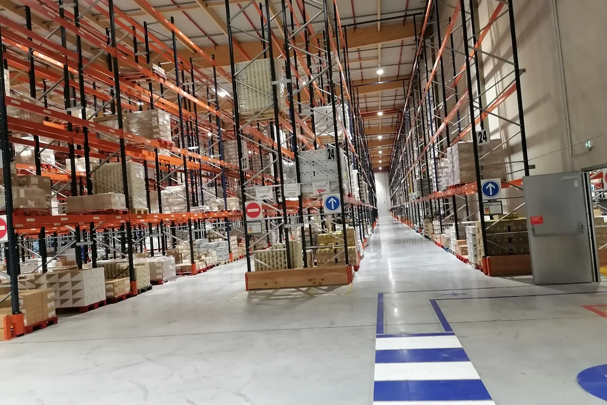 Pasillo de un almacén con estanterías de palés, ubicaciones numeradas y marcas de circulación en el suelo